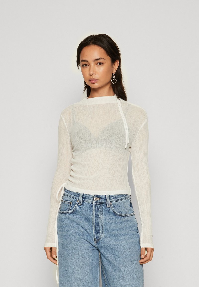 Polo alb, texturat, cu mânecă lungă, cu legături pe lateral; textură transparentă peste un sutien gri deschis. Asortat cu blugi din denim albastru cu talie înaltă.