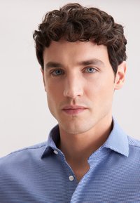 Un giovane con capelli ricci scuri e occhi blu che indossa una camicia a maniche lunghe blu a fantasia su uno sfondo semplice.