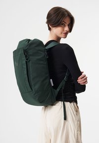 Grüner Stoffrucksack mit zwei vorderen Reißverschlüssen, verstellbaren Trägern und abgerundetem oberen Teil. Minimalistisches Design mit roten und grünen Akzentpunkten.