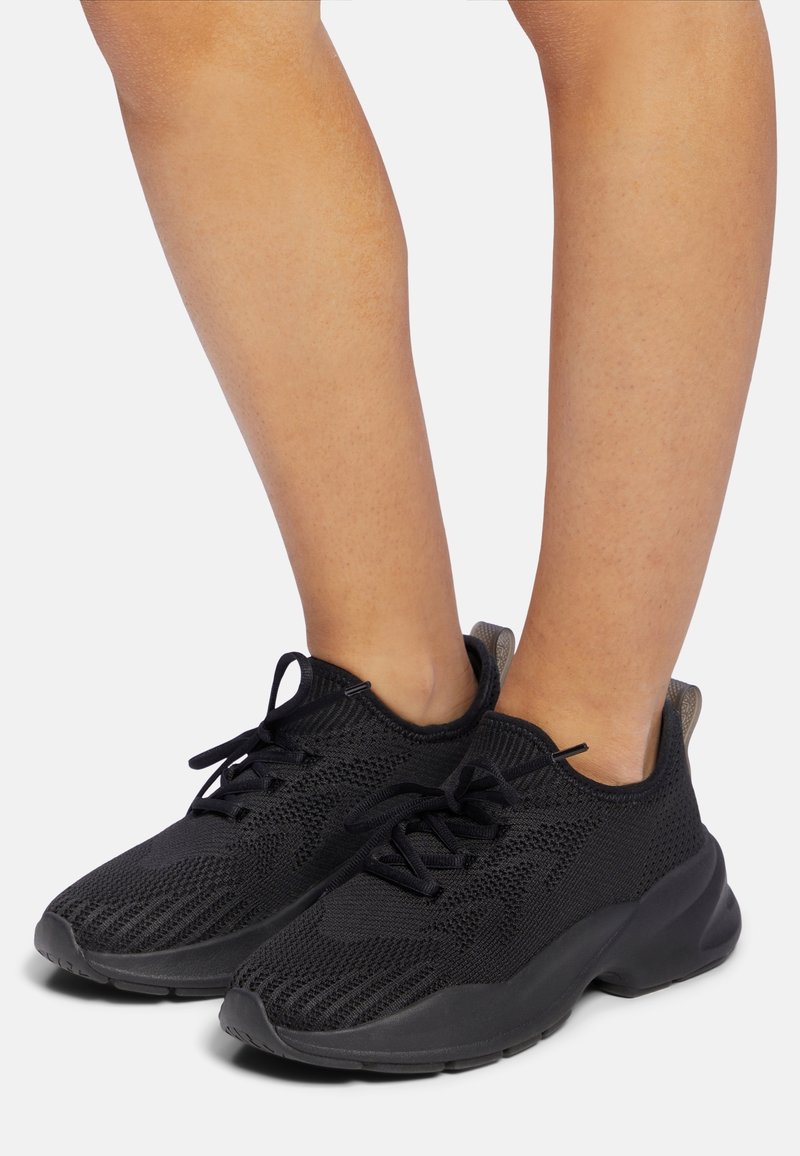 Personne portant des baskets en tissu tricoté noir avec des lacets noirs et des semelles en caoutchouc épaisses sur fond clair.