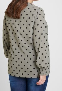 Blouse verte à pois noirs, à manches longues, avec des poignets froncés et une coupe décontractée, associée à un jean bleu. Le tissu semble lisse.