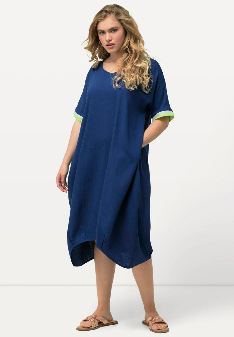Marineblauwe oversized jurk met korte mouwen, voorzien van groene accenten, een ronde halslijn, zijzakken en een asymmetrische zoom. Zachte stofstructuur.