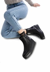 Botas de tobillo de cuero negro con suela de plataforma, con cremallera lateral y detalles texturizados. Llevadas con jeans de un azul claro y un calcetín de red.