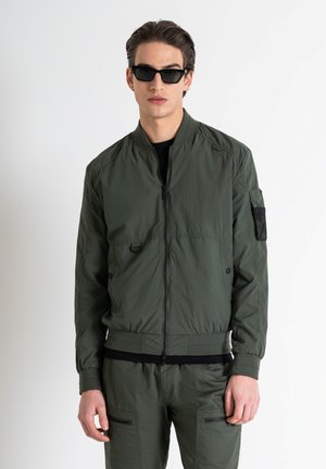 Olive green bomber jacket made of lightweight fabric with a front zipper, ribbed cuffs, and a mesh pocket detail on the left sleeve.