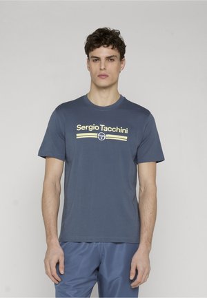 Giovane uomo che indossa una t-shirt blu Sergio Tacchini con logo giallo e pantaloni blu, in piedi davanti a uno sfondo chiaro e uniforme.