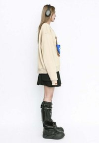 Sudadera oversized beige, falda mini negra y botas de goma negras hasta la rodilla. Los accesorios incluyen un bolso azul. Cabello largo, llevando auriculares.