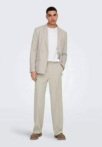 Man wearing beige suit with white t-shirt and brown shoes, standing with one hand in pocket against plain background.