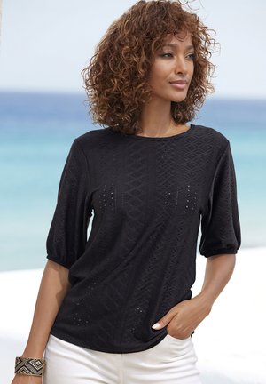 Schwarzer Pullover mit diamantförmigem Muster, kurzen Puffärmeln und einem Schlüsselloch-Ausschnitt. Kombiniert mit hellen Hosen.