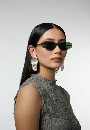 Femme aux cheveux noirs lisses porte des lunettes de soleil fines vert foncé, de grandes boucles d'oreilles argentées abstraites et un haut gris sans manches texturé, regardant vers la droite.