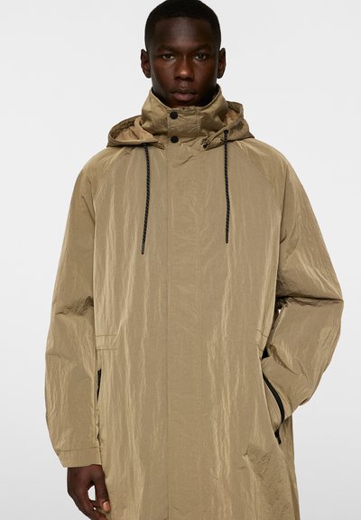 Beige lång jacka med en strukturerad yta, justerbar huva och svarta dragsnören. Har sidofickor och en dragkedja på framsidan.