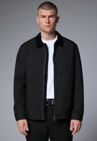Chaqueta negra con cremallera y cuello suave, con un bolsillo en el pecho izquierdo, llevada sobre una camiseta blanca, combinada con pantalones oscuros. Textura suave.
