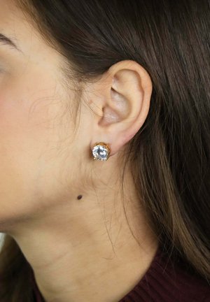 Linkeroor van een vrouw met lang bruin haar naar achteren, met een gouden oorknopje met een ronde heldere edelsteen, zichtbare huidvlek onder het oor.