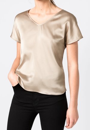 Femme portant un chemisier en satin beige à manches courtes avec col en V et un pantalon noir, une main posée négligemment dans sa poche.