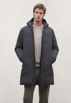 Joven con abrigo de invierno oscuro con capucha sobre un suéter gris claro y pantalones verde oliva, de pie con las manos en los bolsillos.