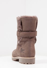 Botín marrón de cuero con forro de pelo sintético, correa ajustable, superficie texturizada y suela de goma. Logo visible en el lateral.