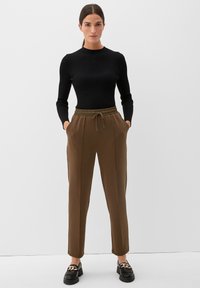 Zwart geribbeld longsleeve shirt gecombineerd met bruine tapered broek met elastische tailleband en zijzakken. Zwarte instappers met kettingdetail.