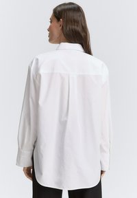 Camicia bianca con colletto a punta, maniche lunghe e piega sul retro. Presenta un orlo curvo e polsini abbottonati.