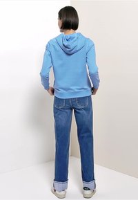 Felpa con cappuccio azzurro chiaro con tasca a marsupio e cappuccio regolabile, abbinata a jeans blu larghi e sneakers bianche con logo.