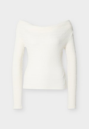 Haut blanc à manches longues, épaules dénudées, texture avec un motif ondulé et silhouette ajustée.