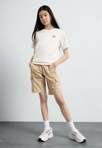 T-shirt en coton blanc avec logo noir, short cargo beige avec poches, baskets blanches avec accents noirs et gris, debout sur un fond gris.