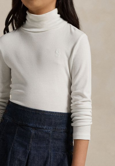 Chica con un suéter de cuello alto blanco y una falda plisada azul oscuro de talle alto con detalles de botones en el lado.