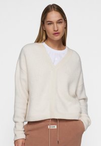 Crème v-hals trui met een zachte textuur en een losse pasvorm. Kenmerkt ribgebreide manchetten en zoom, gelaagd over een witte top. Gepaarde met een bruine joggingbroek.