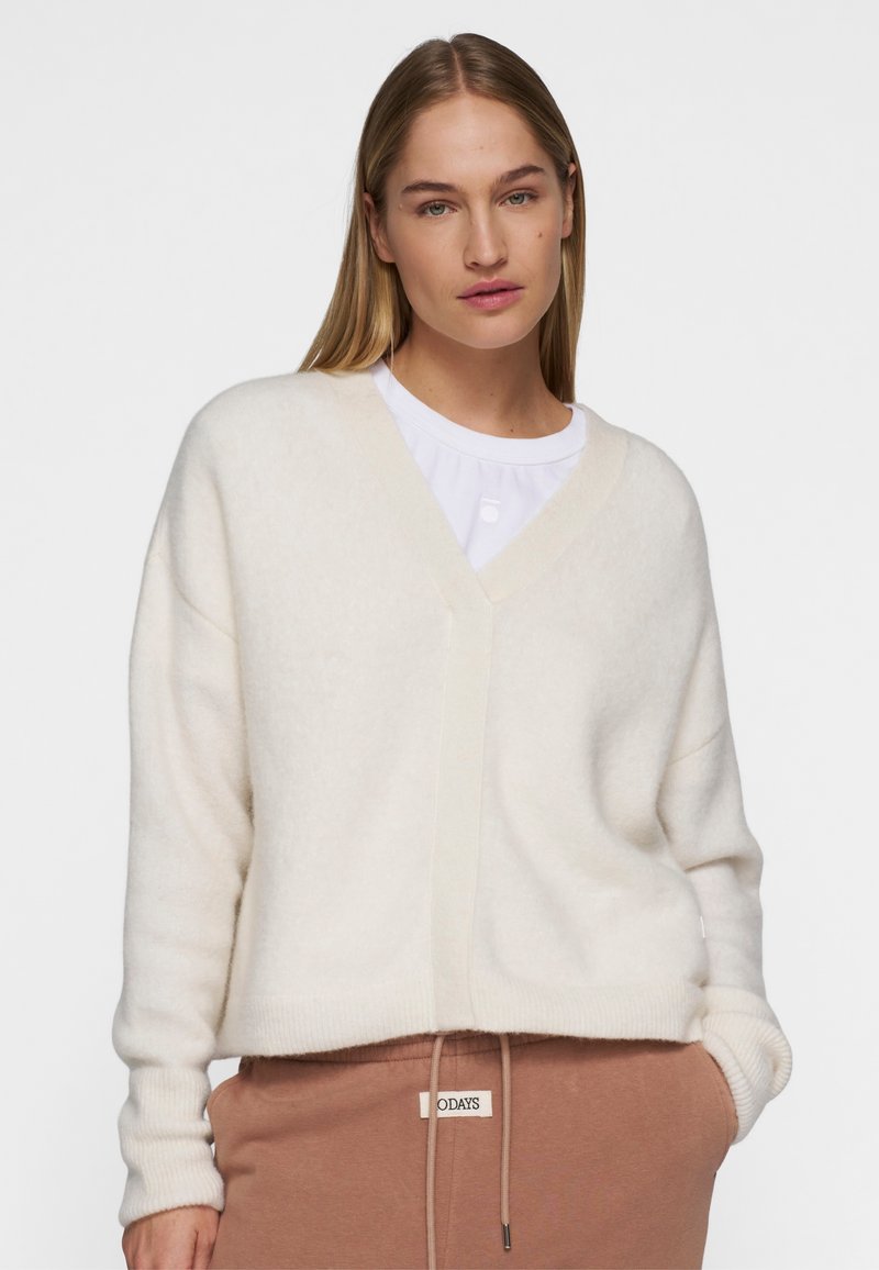 Creme v-hals sweater med en blød tekstur og løs pasform. Har ribbede manchetter og kant, lagdelt over en hvid top. Sæt sammen med brune sweatpants.
