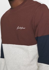 Långärmad sweatshirt i brun, grå och marinblå; har rundad halsringning och vitt logo på bröstet; slät bomullsmaterial.