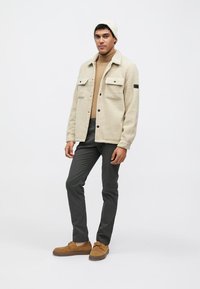 Beige fleecejack met twee voorzakken, gedragen over een beige coltrui, gecombineerd met donkere grijze broek en bruine schoenen, en een witte gebreide muts.