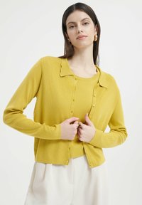 Gelbes Cardigan mit einer gewellten Kragen, gerippter Textur und Knopfverschluss. Trägt über einem passenden gelben Oberteil, das lange Ärmel und eine taillierte Passform hat.