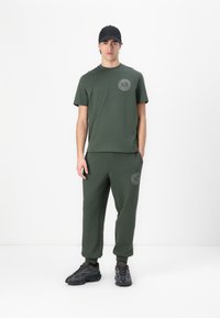 Conjunto de camiseta verde oliva y pantalones de chándal, con un logo redondo en el pecho y en la pierna izquierda. Una gorra negra y zapatillas negras con textura completan el atuendo.
