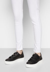 Svarta lädersneakers med tre kardborreband, vit texturerad sula, tillsammans med åtsittande vita jeans. Enkla, moderna linjer.