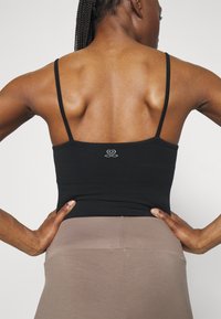 Black tank top with thin straps, featuring a fitted design. The back showcases an embroidered logo above the waist, paired with beige leggings.