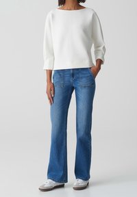 Pull blanc texturé avec un large décolleté, associé à un jean évasé bleu et des baskets aux accents gris. Design simple et décontracté.