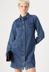 Denimskjolklänning i mörkblått med långa ärmar, knapplåsning, två bröstfickor och en något rundad nederkant. Slät textur.