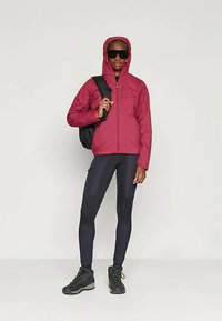 Bordo izolirana jakna s kapuljačom, crne sunčane naočale, crni ruksak, uske crne tajice s teksturiranim detaljima i crne planinarske čizme.