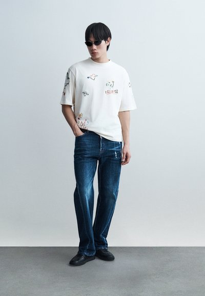 Νεαρός άνδρας με γυαλιά ηλίου, λευκό μπλουζάκι με στάμπα, μπλε τζιν και μαύρα παπούτσια, στέκεται μπροστά σε ένας απλό ανοιχτό γκρι τοίχο.