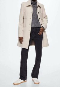 Beige uldfrakke med fire forlommer, grå turtleneck, sorte jeans og hvide sneakers med en gummisål.