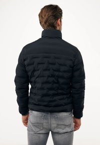 Černá péřová bunda s vysokým límcem, texturovaným prošívaným designem a elastickými manžetami. Nositá s světle šedými džíny, viděno ze zadní strany.