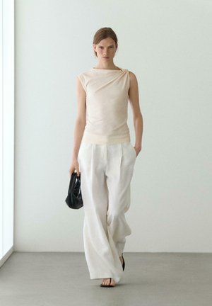 Femme marchant en haut beige sans manches et pantalon blanc large, tenant un petit sac à main noir, devant un mur blanc uni.