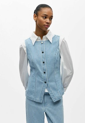 Frau trägt eine hellblaue Weste mit vertikalen Streifen und passende Hose über einem weißen Hemd mit Kragen, schaut nach rechts mit den Händen hinter dem Rücken.
