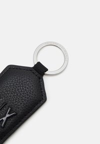 Schwarzer Leder-Schlüsselanhänger in achteckiger Form, mit geprägtem Logo, einer strukturierten Oberfläche und einem silbernen Metallring mit graviertem Markennamen.