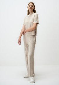 Beige gebreide top met korte mouwen en hoge hals, gecombineerd met bijpassende wijde broek. Model draagt witte sneakers. Minimalistisch ontwerp en zachte textuur.