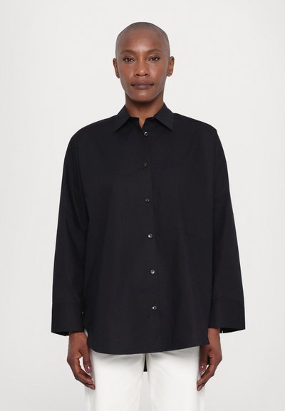 Chemise noire oversize avec col pointu, manches longues, fermeture à boutons sur le devant et poche poitrine, fabriquée en tissu lisse.