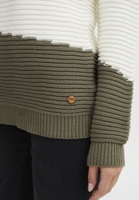 Stickad tröja med horisontella randningar i grädde och olivgrön, med strukturerad ribbning, råa kanter och en accent av trädetalj som logo.