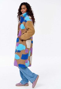 Kobieta uśmiechająca się, ubrana w długi beżowy polarowy płaszcz w niebieskie, fioletowe i białe łatki, niebieskie dżinsy i różowe trampki, stojąca z boku.