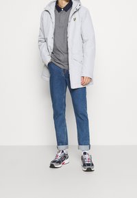Hellgraue wasserdichte Jacke mit Kapuze, ausgestattet mit einem Reißverschluss und Logo, getragen über einem grauen Poloshirt und blauen hochgekrempelten Jeans, kombiniert mit Sneakers.