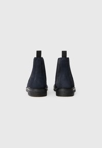 Une paire de bottes Chelsea en daim bleu marine foncé vues de l'arrière, montrant des tirettes noires et des semelles robustes noires sur un fond blanc.