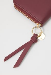Burgundy leather wallet with gold zipper, ring, and a tassel accent. Smooth texture with a sleek rectangular shape.
