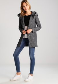 Manteau gris à capuche avec une texture douce, fermeture éclair sur le devant et deux poches, associé à un haut noir et un jean bleu, avec des baskets blanches.